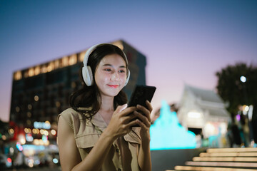 A cheerful woman listening to music with headphones while holding her phone in a lively city at dusk, surrounded by colorful lights and urban atmosphere.