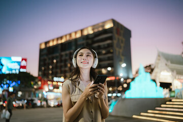 A cheerful woman listening to music with headphones while holding her phone in a lively city at dusk, surrounded by colorful lights and urban atmosphere.