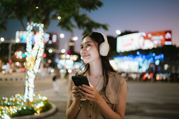 A cheerful woman listening to music with headphones while holding her phone in a lively city at dusk, surrounded by colorful lights and urban atmosphere.