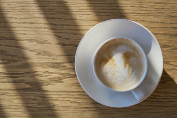 Coffee cup with latte art foam sitting on wooden table with sun shadows