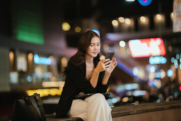 A smiling woman using her smartphone at night in a lively city setting, creating a warm and relaxed atmosphere with colorful blurred lights in the background.