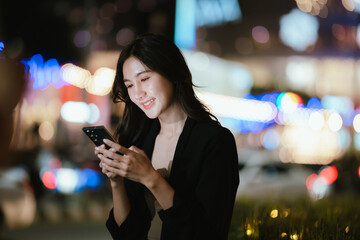 A smiling woman using her smartphone at night in a lively city setting, creating a warm and relaxed atmosphere with colorful blurred lights in the background.