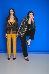 Fototapeta premium Young beautiful twin sisters showcase stylish attire against a vibrant blue backdrop