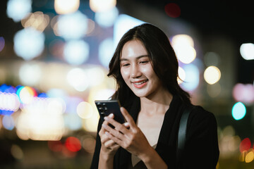 A smiling woman using her smartphone at night in a lively city setting, creating a warm and relaxed atmosphere with colorful blurred lights in the background.