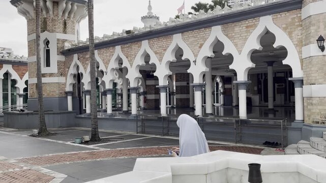 Anonymous woman in hijab sits looking at cellphone peacefully outside Masjid Jamek Sultan Abdul Samad in Kuala Lumpur, with its distinctive Moorish arches and brick walls in the background