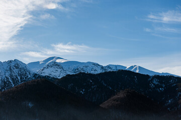 Majestic snow-capped mountain ridge under a vibrant blue sky and sweeping clouds. Dark, forested foothills lead up to the illuminated, icy summits