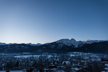Snow-covered Zakopane nestled beneath the majestic, jagged, and dark peaks of the Tatra Mountains. A quiet, cold winter landscape at dawn, dusk