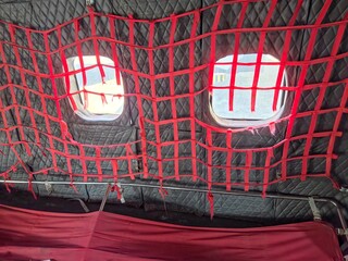 Interior view looking up at the ceiling of an aircraft or structure, showing black quilted insulation and two oblong windows covered by a tight, red cargo or safety net.