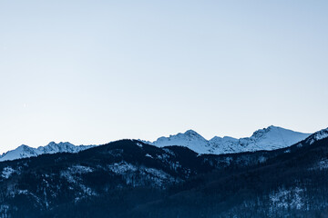 Serene winter mountain skyline with snow-covered peaks rising above dark, densely forested lower slopes under a pale, cold, gradient sky. Zakopane, Poland