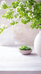 A still life image featuring white flowers and green leaves in a vase, with a small bowl of green herbs on a white table, natural light.