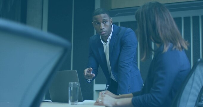 Leaning man in navy suit gesturing and speaking over conference table with open laptop and papers
