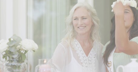 Smiling senior woman sitting in lace blouse at dining table, friend adjusting floral crown and sash