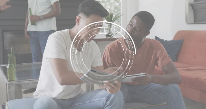 Collaborating men wearing white tee, rust tee leaning over tablet in living room, HUD, green bottle
