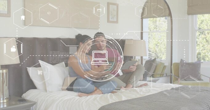 Using smartphone and tablet, couple wearing casual clothes sitting on king-size bed, HUD graphics