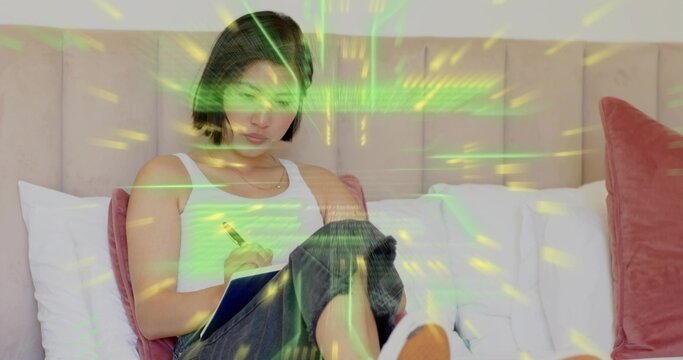 Writing woman sitting on bed wearing tank top holding blue notebook, pen, lit by green projection