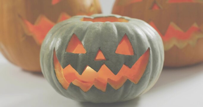 Fototapeta Glowing gray-green carved pumpkin lying on white tabletop, with two orange pumpkins lit, ridged