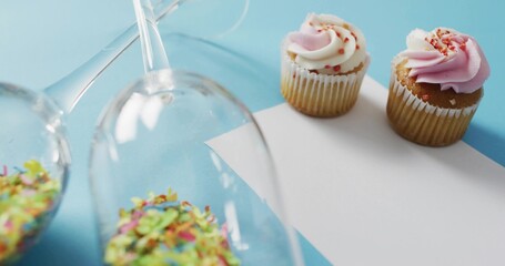 Showing two frosted cupcakes on card at pale blue surface, glasses spilling sprinkles, copy space © vectorfusionart
