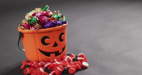 Showing orange jack o'lantern bucket spilling foil chocolates, gummies on gray backdrop, copy space