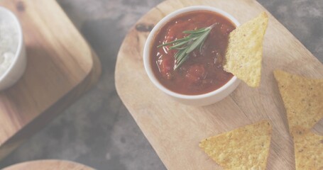 Showing white bowl of chunky tomato salsa with rosemary on wooden board, dark slate, nachos, salt