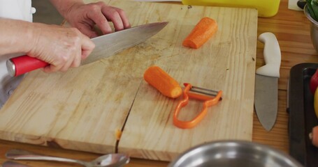 Slicing senior hands holding red knife on cracked board in kitchen, white sleeve visible
