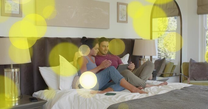 Sitting couple in casual clothes leaning on dark headboard in master bedroom, viewing tablet