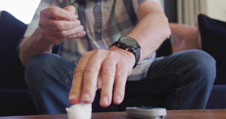 Handling lancet, senior male in plaid shirt, on sofa reaching cotton ball on table with glucometer