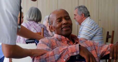 Obraz premium Leaning back senior man in wheelchair, staff in scrubs placing hand on shoulder in common room