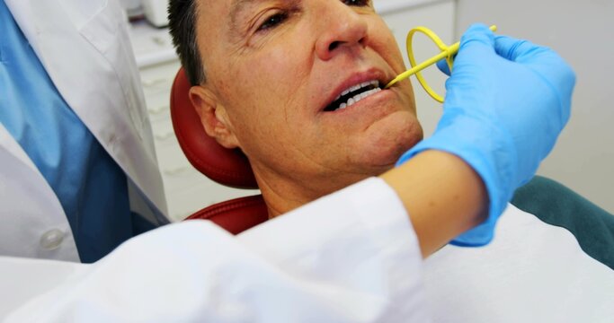 Reclining male patient opening mouth in clinic with yellow suction, blue-gloved white-coat dentist