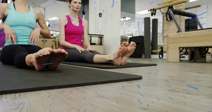 Sitting two women holding legs extended on mats in pilates studio, wearing teal and pink tanks