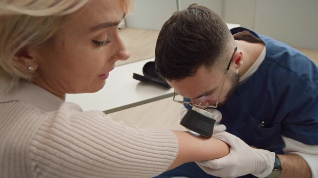 Handheld shot of young male doctor in scrubs examining elbow using dermatoscope while middle-aged Caucasian female patient explaining symptoms during medical appointment in clinic
