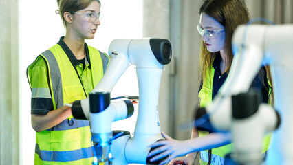 Two female engineers in safety vests and goggles collaborate in a tech lab. They are inspecting and programming a high tech robotic arm for a new project.