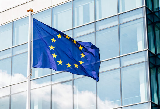 The european union flag with yellow stars on a blue background waves against a modern glass building facade under a clear sky suggesting a formal and sleek urban setting
