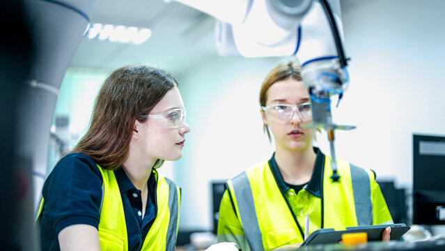 Two female engineers in safety vests collaborate in a tech lab. One woman holds a tablet while the other thinks, looking at a high tech robotic arm.