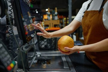 The female cashier is scanning an orange at the supermarket checkout