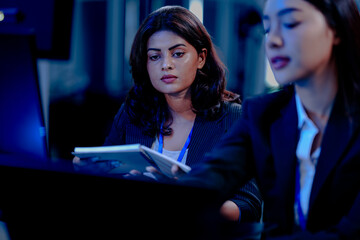 Intense late night focus: Two diverse female professionals collaborate closely, deeply engaged in reviewing critical project data displayed on a computer screen in a modern hub.
