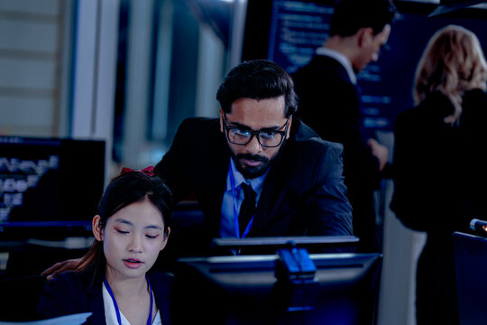 Intense collaboration hub Diverse AI team deep dive into project data late night. Manager guides Asian colleagues reviewing analytics for a new service launch in tech office.