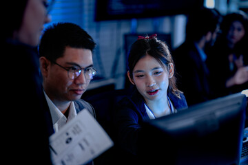 Intense late night project review. Female team leader points out critical data on screen discussing report charts with colleagues in modern high tech operations center.
