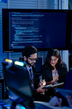 An AI research team brainstorms late night. Innovators discuss machine learning model architecture using a tablet while reviewing training data code displayed on a large screen.