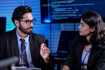 Focused late night strategy session. Male manager with glasses gestures explaining complex points to attentive female colleague in modern high tech command center office.
