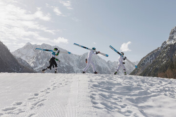 Athletes in white outfits walk on a snow-covered slope, holding skis, ready for ski jumping in a mountain setting.