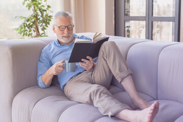 Old elderly senior handsome man with grey hair reading a book lying and relaxing with cup mug of tea coffee on the sofa in the living room at home . Social distance and isolation concept