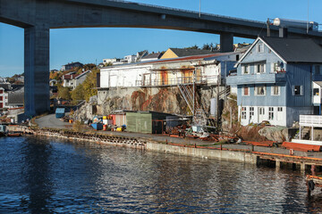 A rugged waterfront scene beneath a sweeping bridge shows weathered houses