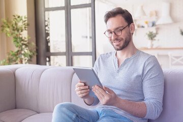 Confident young caucasian man using digital tablet sitting on the sofa, relaxing and reading e-book, e-learning, checking social media, doing online shopping, have zoom call conference at home indoors