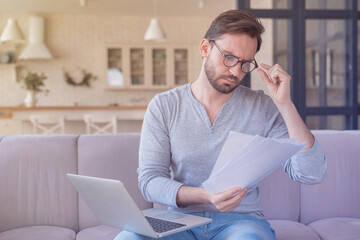 Serious young caucasian man businessman freelancer entrepreneur investor trader paying bills, checking documents and doing paperwork using laptop at home indoors. Remote work freelance concept