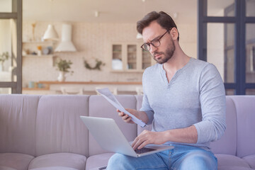 Concentrated serious young caucasian man businessman freelancer paying domestic bills, counting money, doing paperwork using laptop at home. Home office and occupation. Remote work concept