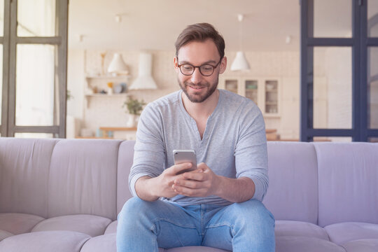Relaxed young caucasian man in casual clothes wearing eyeglasses using smartphone for reading news online, checking social media, loading feed, e-learning and working at home indoors - Powered by Adobe