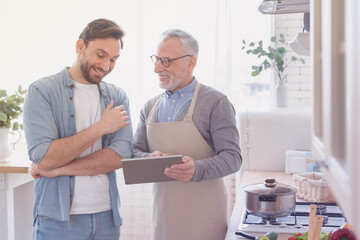 Young caucasian man son helping his old mature senior father with cooking searching recipe online using digital tablet. Happy father`s day! I love you, dad! Men cooking together