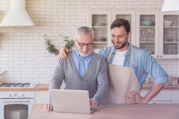 Cheerful young adult son teaching his old senior elderly father how to search recipes online using internet laptop in the kitchen while cooking food together. Happy father`s day!
