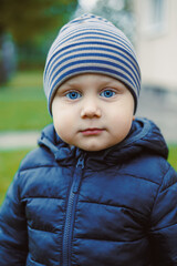 Bright-eyed child in a cozy blue jacket and striped hat enjoys a chilly day outdoors, surrounded by greenery and gentle sunlight