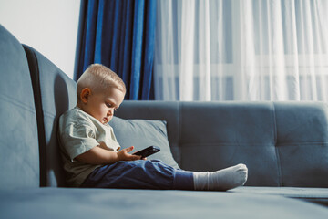 Young child focused on smartphone while sitting on a cozy blue couch in a bright living room during a quiet afternoon